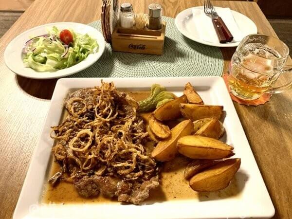 A close-up, high-angle shot on a wooden table in an Austrian restaurant: a hearty plate of Zwiebelrostbraten features a tender beef steak topped with a generous layer of crispy, golden-brown fried onions. Next to it sits a rounded glass mug (Krügerl) of Murauer Märzen on tap, already more than half-empty, with only a small amount of pale golden beer and foam remaining.