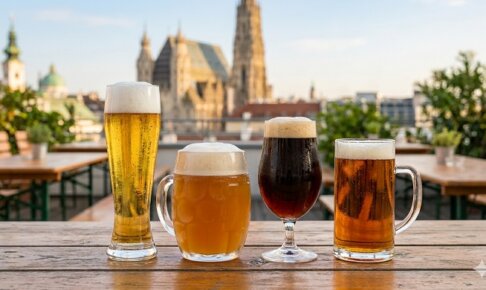 A wide-angle, cinematic shot of four different types of Austrian beers in various glasses—including a pilsner flute, a dimpled mug, and a tulip glass—standing on a rustic wooden outdoor table. Each beer shows a different hue from pale gold to dark amber, topped with fresh white foam and condensation on the glass. The background features a blurred, sunlit view of Vienna's historic skyline, including the spire of St. Stephen's Cathedral.