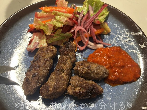 青いお皿に盛り付けられた3本のチェヴァピ、生野菜サラダ、そしてたっぷりの赤いアイヴァルソース。